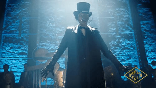 A woman with a fake moustache, black top hat, and black trenchcoat on stage about to wow everyone with their performance