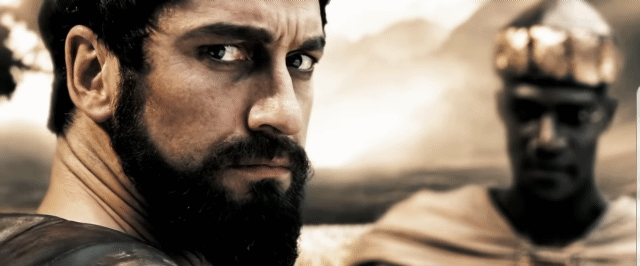 A black haired white man with a long black beard in Sparta looking at his long dark haired wife nodding her head. Behind the dark haired white man stands a black ambassador from Persian in a stylized environment