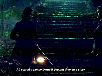 A darkly light screen grab of a Japanese woman with her black hair in a tight bun and wearing a black coat hoping a rifle pointed downward talks to a white man with wavy brown hair and a scruff in a thick jacket. Behind them are wet and slimmer stairs that led into a cave. The woman is holding a lantern, the only light source. On the bottom of the image there is text that says: All sorrows can be borne if you put them in a story. 